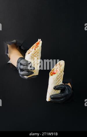 Hände, die Döner oder Shawarma durch gerissene Löcher auf schwarzem Papierhintergrund halten. Street-Food-Konzept Stockfoto
