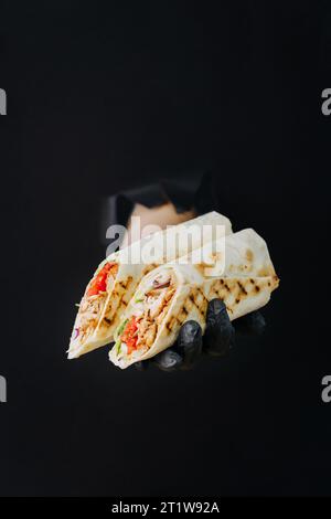 Hand hält Döner oder Shawarma durch gerissene Löcher auf schwarzem Papierhintergrund. Street-Food-Konzept Stockfoto