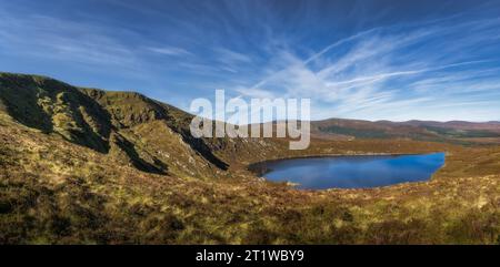 Breites Panorama mit rauem Hügel des Tonelagee Mountain und herzförmigem See, Lough Ouler, reflektierendem blauen Himmel. Wandern in den Wicklow Mountains, Irland Stockfoto