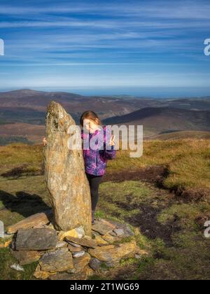 Ein junges Mädchen mit einem kleinen Obelisken auf der Spitze des Tonelagee-Berges mit einer Bergkette im Hintergrund. Wandern in den Wicklow Mountains, Irland Stockfoto