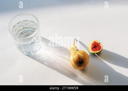 Ein Glas mit Wasser im Sonnenlicht, Birne und der Hälfte Feigenfrucht auf weißem Tisch. Stillleben. Stockfoto