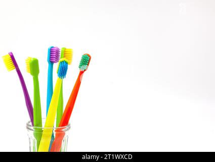 Drei Zahnbürsten – violett, orange und gelb mit verschiedenfarbigen Borsten in einer klaren Tasse auf weißem Hintergrund. Isoliert. Vertikal. Foto Stockfoto