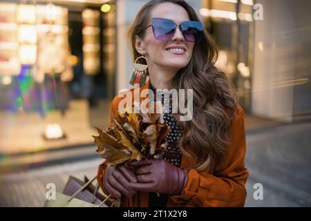 Hallo oktober. Glückliche, moderne 40-jährige Frau in orangefarbenem Trenchcoat mit Einkaufstaschen und herbstgelben Blättern in der Nähe eines Shops in der Stadt. Stockfoto