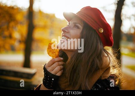 Hallo Herbst. Glückliche moderne 40-jährige Frau mit rotem Hut mit Herbstblatt, Schal und Handschuhen, die auf einer Bank im Stadtpark sitzt. Stockfoto