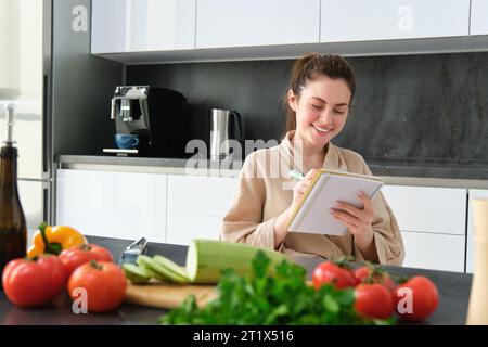 Porträt einer glücklichen jungen Frau schreibt Menü zum Abendessen auf, sitzt in der Küche neben Gemüse, macht Lebensmittellisten zum Einkaufen, posiert im Bademantel Stockfoto