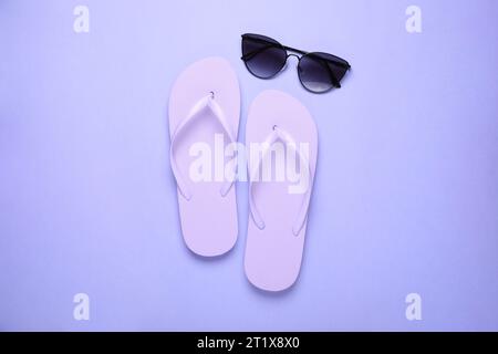 Stilvolle Flip-Flops und Sonnenbrille auf hellviolettem Hintergrund, flach geschnitten Stockfoto