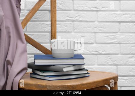 Keramikbecher und Stapel Bücher auf Holzstuhl vor weißer Ziegelwand, Platz für Text Stockfoto