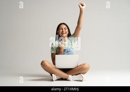 Glückliches lächelndes Teenager-Mädchen, das auf grauem Hintergrund sitzt, mit Laptop auf den Beinen. Stockfoto