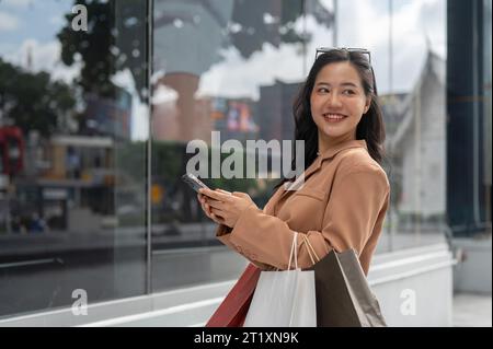 Eine attraktive und lächelnde asiatische Frau in trendigen Kleidern, die ihr Smartphone und ihre Einkaufstaschen hält und ihren Einkaufstag in der Stadt genießt. Städtisches Leben Stockfoto