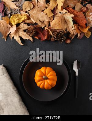 Kürbissuppe-Konzept. Festlicher Herbsttisch mit Herbstblättern und Herbstkürbis in einer schwarzen Suppenschüssel auf schwarzem Hintergrund. Draufsicht. Stockfoto