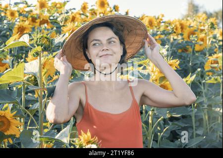 Schöne kaukasische Frau in Sonnenblumen, Porträt einer Frau mit vietnamesischem Hut, die bei Sonnenuntergang durch ein blühendes Sonnenblumenfeld geht. Frieden, Stockfoto