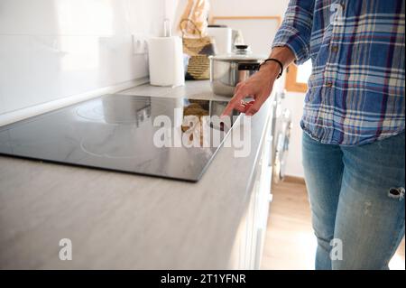 Nahaufnahme eines Mannes, der die Hand auf der schwarzen Glasoberfläche eines Elektroherds hält, ihn anschaltet und das Programm anpasst, während er das Abendessen in einem Mini kocht Stockfoto