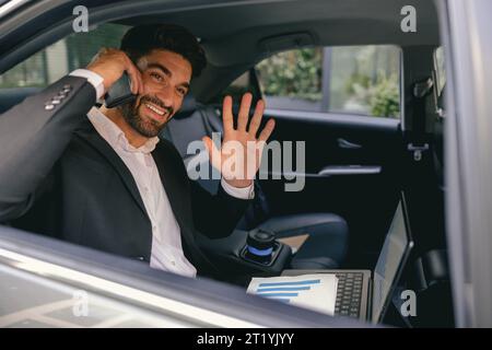 Lächelnder Geschäftsmann im Anzug mit Laptop, der telefoniert, sitzt im Taxi und winkt Hi im offenen Fenster Stockfoto