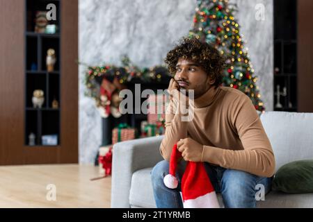 Gelangweilter und trauriger Mann, der zu weihnachten in der Nähe des weihnachtsbaums auf dem Sofa sitzt, hispanic deprimiert im Wohnzimmer einsam keine Freunde für Neujahrsfeier. Stockfoto