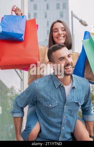Ein Mann, der seine Freundin auf dem Rücken trägt und Spaß mit Einkaufstaschen hat Stockfoto