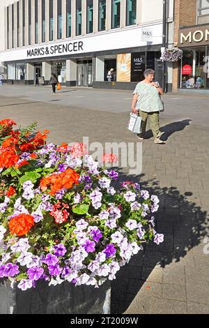 Model veröffentlichte reife Käuferin, die breite Einkaufsmöglichkeiten auf der High Street Straßenbelag bunter Blumenkäufer Marks & Spencer Store Brentwood Essex England spaziert Stockfoto