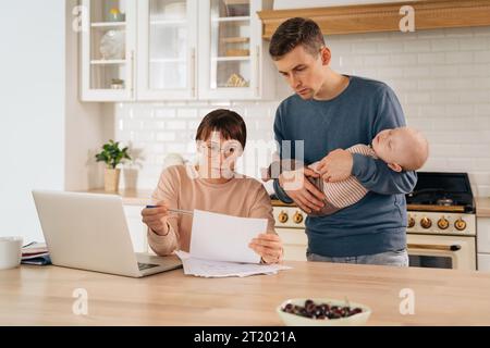 Business und Familie. Millennials Eltern mit schlafendem Baby Sohn, der in der Küche am Laptop arbeitet, an Plänen oder finanziellen Problemen denkt, Hausrechnungen bezahlt, Familienaufgaben und -Abschlüsse diskutiert Stockfoto