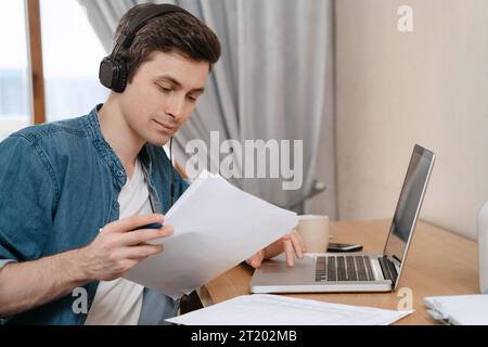 Zielgerichteter junger Schüler, der Kopfhörer trägt, mit einem Laptop für Fernunterricht in seinem Zimmer, Hören von Audiokursen, Fremdsprachenlernen, Online-Webinar ansehen, elektronisches Lernkonzept Stockfoto