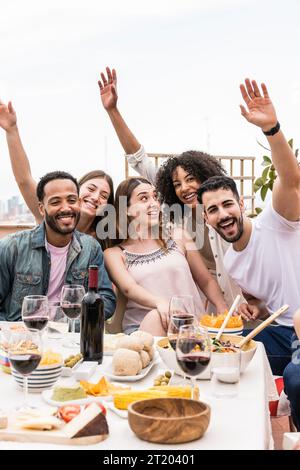 Fröhliche multirassische Leute lachen auf einer Dachparty. Eine positive Gruppe von Freunden, die sich beim Abendessen auf einer Terrasse amüsieren. Stockfoto