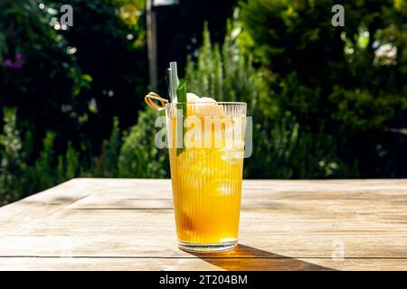 Frischer hausgemachter Lychee-Eistee mit Pandanblättern, serviert auf Holztisch im Freien. Sommer kaltes Obstgetränk am sonnigen Nachmittag mit grünem Garten dahinter Stockfoto