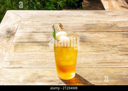 Frischer hausgemachter Lychee-Eistee mit Pandanblättern, serviert auf Holztisch im Freien. Sommer kaltes Obstgetränk am sonnigen Nachmittag mit grünem Garten dahinter Stockfoto