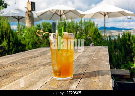 Frischer hausgemachter Lychee-Eistee mit Pandanblättern, serviert auf Holztisch im Freien. Sommer kaltes Obstgetränk am sonnigen Nachmittag mit grünem Garten dahinter Stockfoto