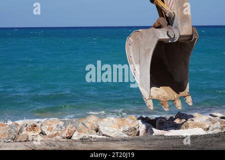 Nahaufnahme einer Löffelschaufel eines Raupenbaggers, der auf der Baustelle arbeitet. Dahinter befindet sich ein Felsenstrand und das Meer von Kreta. Stockfoto