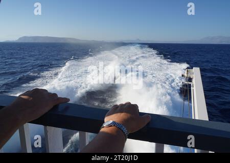 Schiffs-Geländer mit zwei Händen, die darauf gelehnt sind. Im Hintergrund ist Kielwasser hinter dem beweglichen Schiff zurückgelassen. Stockfoto