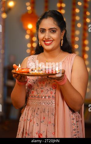 Vertikales Porträt einer glücklichen jungen indischen Frau, die in die Kamera schaut, indem sie die diwali-diya-Lampe zu Hause hält - Konzept der traditionellen Beleuchtung, festlich Stockfoto