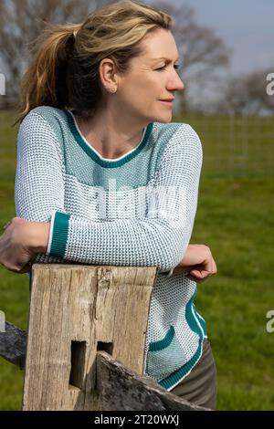 Attraktiv lächelnde glückliche Frau mittleren Alters, die sich auf dem Land auf einem Zaun ruht Stockfoto