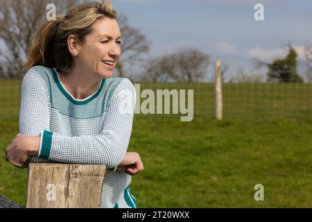 Attraktiv lächelnde glückliche Frau mittleren Alters, die sich auf dem Land auf einem Zaun ruht Stockfoto