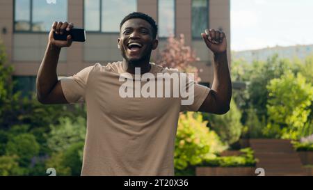 Glücklicher aufgeregter erfolgreicher Afroamerikaner mit Smartphone Erfolg Sieger Mann mit Handy erreichen in der Stadt Glück männlich draußen gewinnen Stockfoto