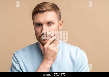 Nachdenklicher ernsthafter bärtiger Mann mittleren Alters, der das Kinn berührt und die Kamera auf beigefarbenem Hintergrund betrachtet Stockfoto