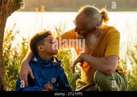 Ein herzerwärmender Moment, als ein liebevoller Großvater und sein kleiner Enkel bei Sonnenuntergang im Gras an einem Waldsee sitzen und eine besondere Bindung an die ruhige Schönheit der Natur teilen. Großvater und Enkel Bonding am Sunset Lake. Hochwertige Fotos Stockfoto