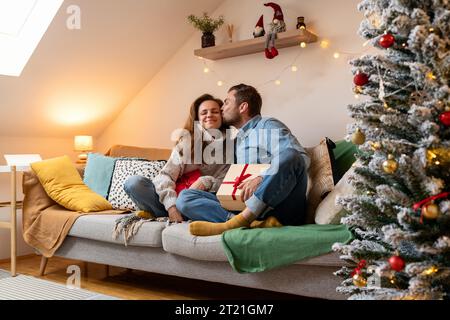Der Mann gibt der Frau ein Weihnachtsgeschenk. Der Freund gibt seiner Freundin Weihnachtsgeschenk und küsst sie auf die Wange. Stockfoto