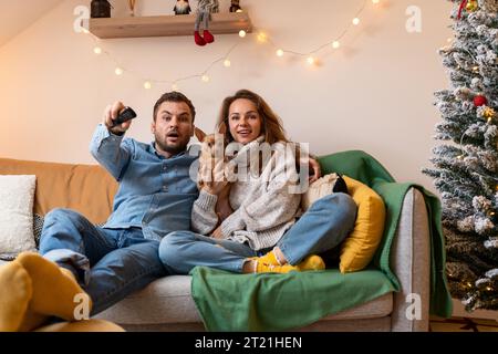 Junge verheiratete Paare und ihr kleiner Schoßhund schauen während der Weihnachtsferien zusammen fern. Verwenden der Fernbedienung. Stockfoto