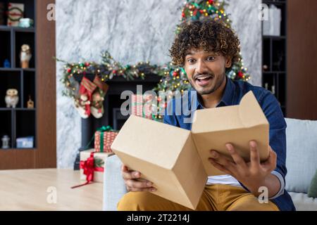 Glücklicher weihnachtsmann, der Neujahrsfeiertage feiert und ein Geschenk auspackt, das von einem Postkurier geschickt wurde, fröhlich lachend hispanic sitzt im Wohnzimmer auf dem Sofa. Stockfoto