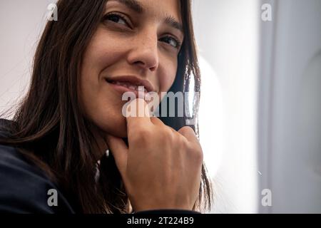 Positive junge Frau mit dunklem Haar, das das Kinn berührt und in hellem Raum daheim wegschaut Stockfoto
