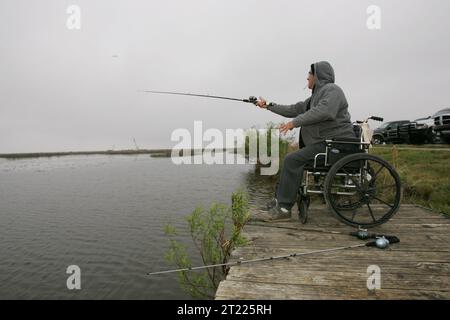 Mann im Rollstuhl, Angeln vom Steinpier an bewölktem Tag. Themen: Fischerei; Freizeit. Lage: Louisiana. Fish and Wildlife Service Site: LACASSINE NATIONAL WILDLIFE REFUGE. Stockfoto