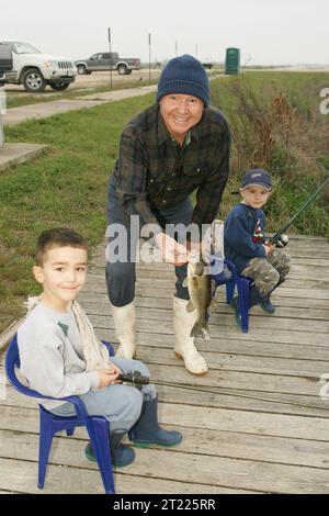 Ein Mann, der stolz Fisch zeigt, der gerade von zwei kleinen Jungs gefangen wurde, die in kleinen Stühlen auf dem Angelsteg saßen. Themen: Kinder, Fischerei, Freizeit. Lage: Louisiana. Fish and Wildlife Service Site: LACASSINE NATIONAL WILDLIFE REFUGE. Stockfoto