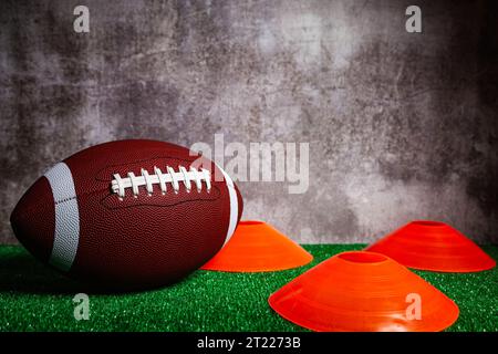 Ein american Football Ball und drei orangefarbene Kegel über grünem Grasboden vor einer grauen Betonmauer. Stockfoto