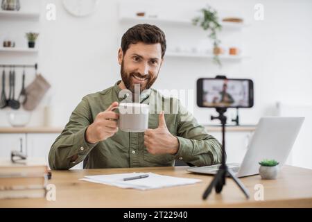 Optimistischer Influencer, der Vlog in der Küche aufzeichnet und ein modernes Smartphone auf einem Stativ verwendet, das den Daumen nach oben zeigt Stockfoto