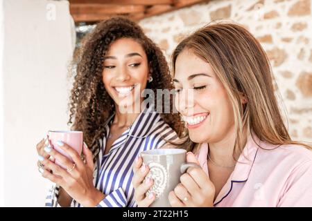 Neue Familiengewohnheiten: Multirassische weibliche Paare trinken eine Tasse Tee auf der Veranda ihres Landhauses - zwei schöne junge Frauen trinken Kaffee A Stockfoto