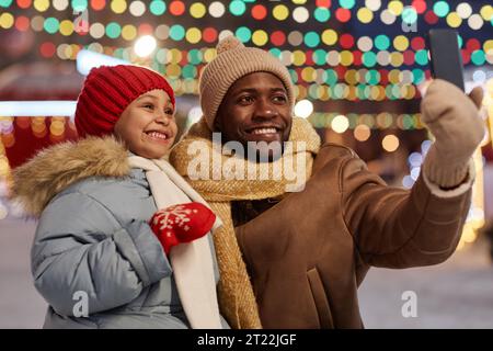 Schwarzer Mann und Tochter machen Selfie-Fotos im Freien zusammen auf dem Weihnachtsmarkt und lächeln, Kopierraum Stockfoto