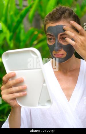 Eine Frau trägt eine graue Tonmaske auf ihr Gesicht und schaut in den Spiegel. Eine Frau in einem Innenhof vor dem Hintergrund von grünem Laub im Stockfoto