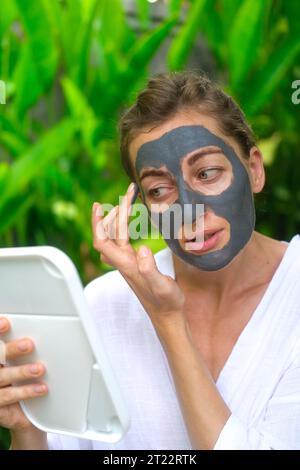 Eine Frau trägt eine graue Tonmaske auf ihr Gesicht und schaut in den Spiegel. Eine Frau in einem Innenhof vor dem Hintergrund von grünem Laub im Stockfoto