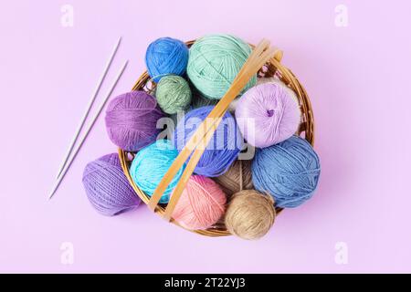 Viele bunte Strickbälle in einem Korb mit Stricknadeln auf einem pastellfarbenen lila Hintergrund. Set für das Hobbys, warme Kleidung oder Spielzeug zu stricken. C Stockfoto