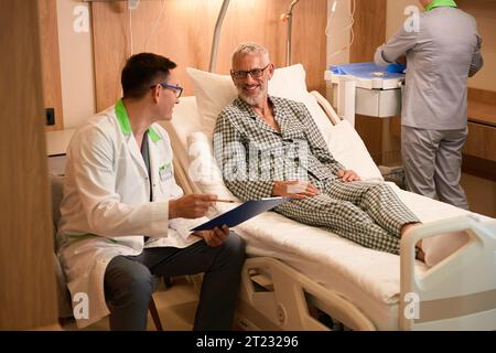 Alter Mann im Pyjama spricht mit behandelndem Arzt im Krankenhauszimmer Stockfoto