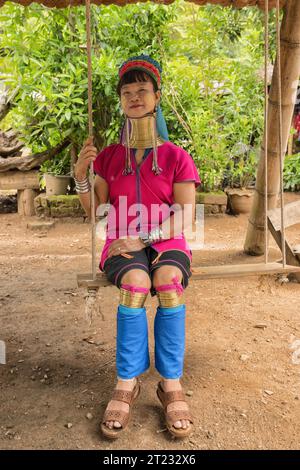Karen Frau aus Pai in Mae Hong Son, Thailand, mit Messing Halsringen sitzt auf einer Holzschaukel im rustikalen Dorf und trägt ein farbenprächtiges Karen Kleid Stockfoto