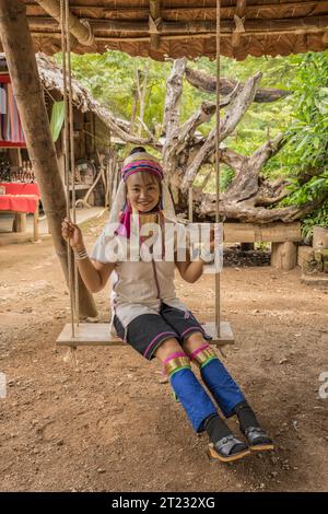 Karen Frau aus Pai in Mae Hong Son, Thailand, mit Messing Halsringen sitzt auf einer Holzschaukel im rustikalen Dorf und trägt ein farbenprächtiges Karen Kleid Stockfoto